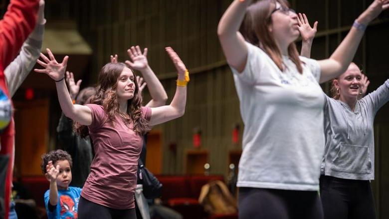 A group of young people dance on a stage. One girl is in focus with her hands waving in the air